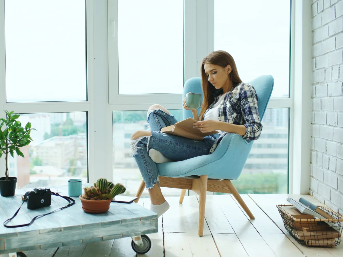 A woman reading in a blue armchair by tall windows overlooking the city